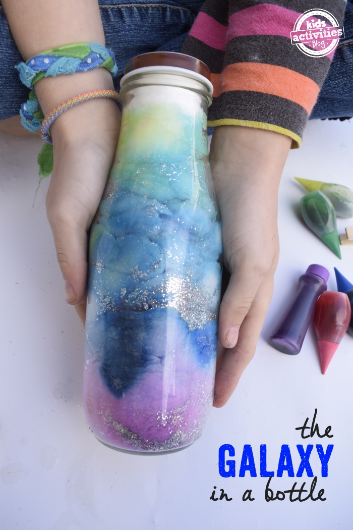 text: DIY Galaxy Jar - Kids Activities Blog - finished glittery galaxy jar held by child on white background