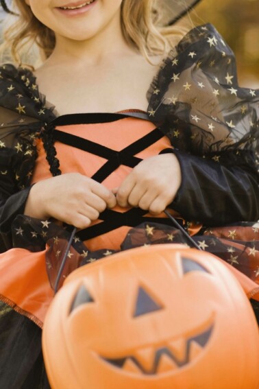 A girl dressed up as a witch is holding an orange plastic pumpkin bucket ready to trick-or-treat in her neighborhood.