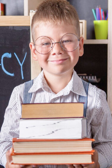 International Literacy Day: Little boy with glasses and a pile of books in his hads, standing in front of a chalkboard - Kids Activities Blog
