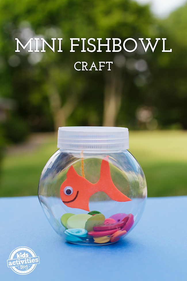 Finished goldfish fish bowl craft for kids sitting on a table outside on a table with a craft paper goldfish hanging inside a small plastic fish bowl - Kids Activities Blog