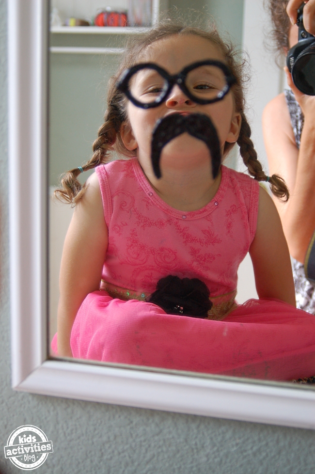 diy mustache and eye glasses clings - little girl looking in mirror with eye glasses on and mustache from diy window clings - Kids Activities Blog