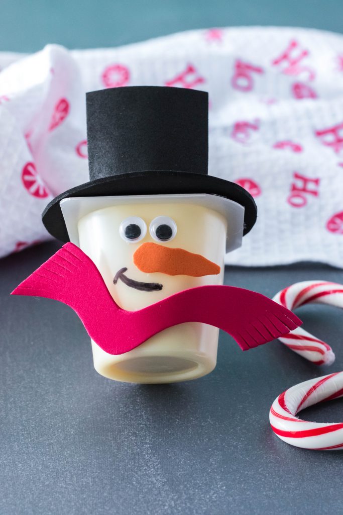 snowman vanilla pudding cup with foam red scarf, orange felt nose, black sharpie smiley face, and googly eyes, and foam black hat