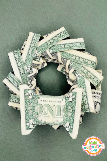 Final result for a dollar bill origami wreath on top of a green background tutorial from kids activities blog